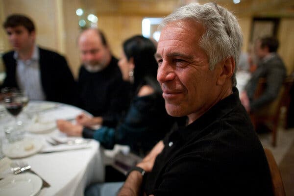 Jeffrey Epstein is shown smiling and wearing a blue shirt as he sits at a dining table. Other people are in the background.